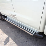 Load image into Gallery viewer, A close-up side view of a white pickup truck showing a silver metal side step bar with black rubber base mounts, installed on the driver's side door. The truck is parked on an unpaved gravel surface.