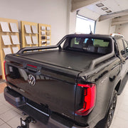 A black Volkswagen Amarok pickup truck with a tonneau cover, photographed from the rear three-quarter angle inside a showroom.