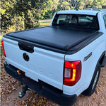 Load image into Gallery viewer, A black tonneau cover installed over the bed of a white Volkswagen Amarok pickup truck, photographed outdoors on a dirt path with trees in the background.