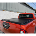 Load image into Gallery viewer, A red Toyota Hilux pickup truck parked outdoors against a white wall and gray metal building, showing a black tonneau cover installed across the bed.
