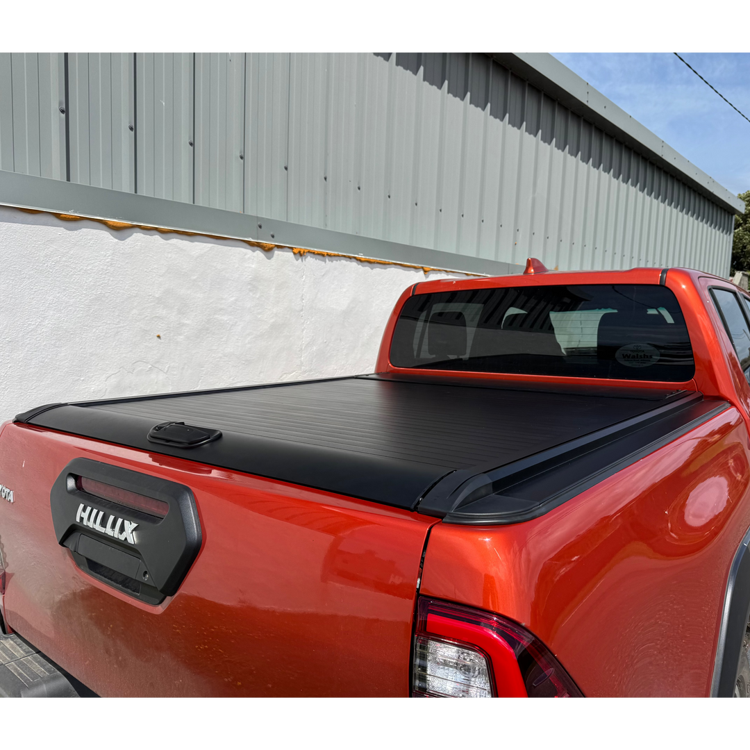 A red Toyota Hilux pickup truck parked outdoors against a white wall and gray metal building, showing a black tonneau cover installed across the bed.