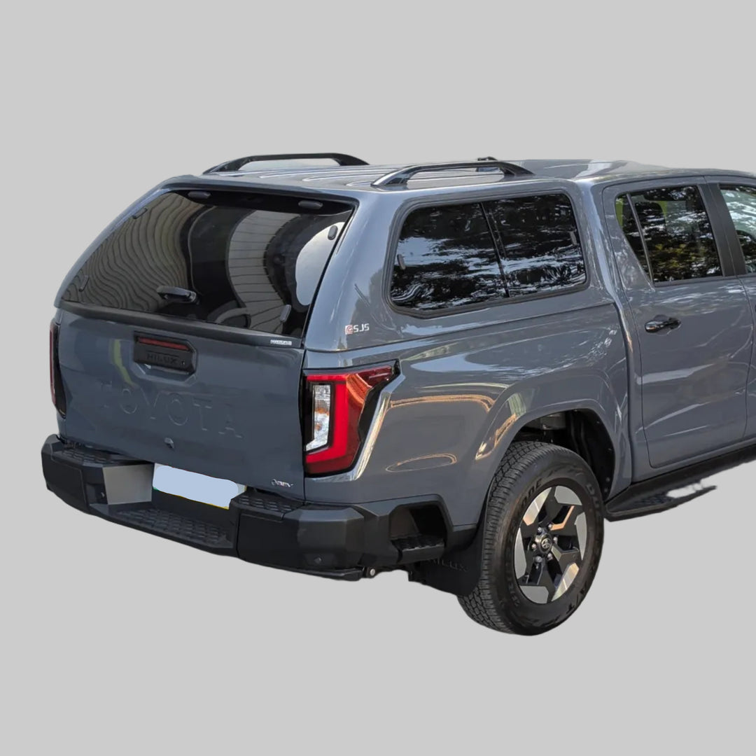 A dark gray pickup truck Travo with a hardtop canopy and black wheels, photographed from the rear three-quarter angle in an outdoor driveway in front of gray garage doors.