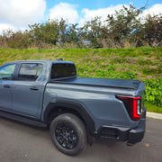 A gray pickup truck parked outdoors on a paved area with a grassy hill and trees in the background. The truck is shown from a rear three-quarter angle, featuring black wheels, a black rear bumper, and a black tonneau-style bed cover.