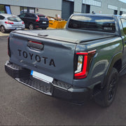 A dark gray Toyota double-cab pickup truck parked outdoors in front of a gray industrial building. The photo shows the rear three-quarter view of the truck