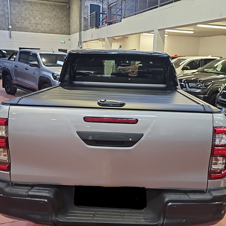 A silver pickup truck Toyota Hilux viewed from the rear in a showroom or garage, with a black tonneau cover roller shutter on the bed 