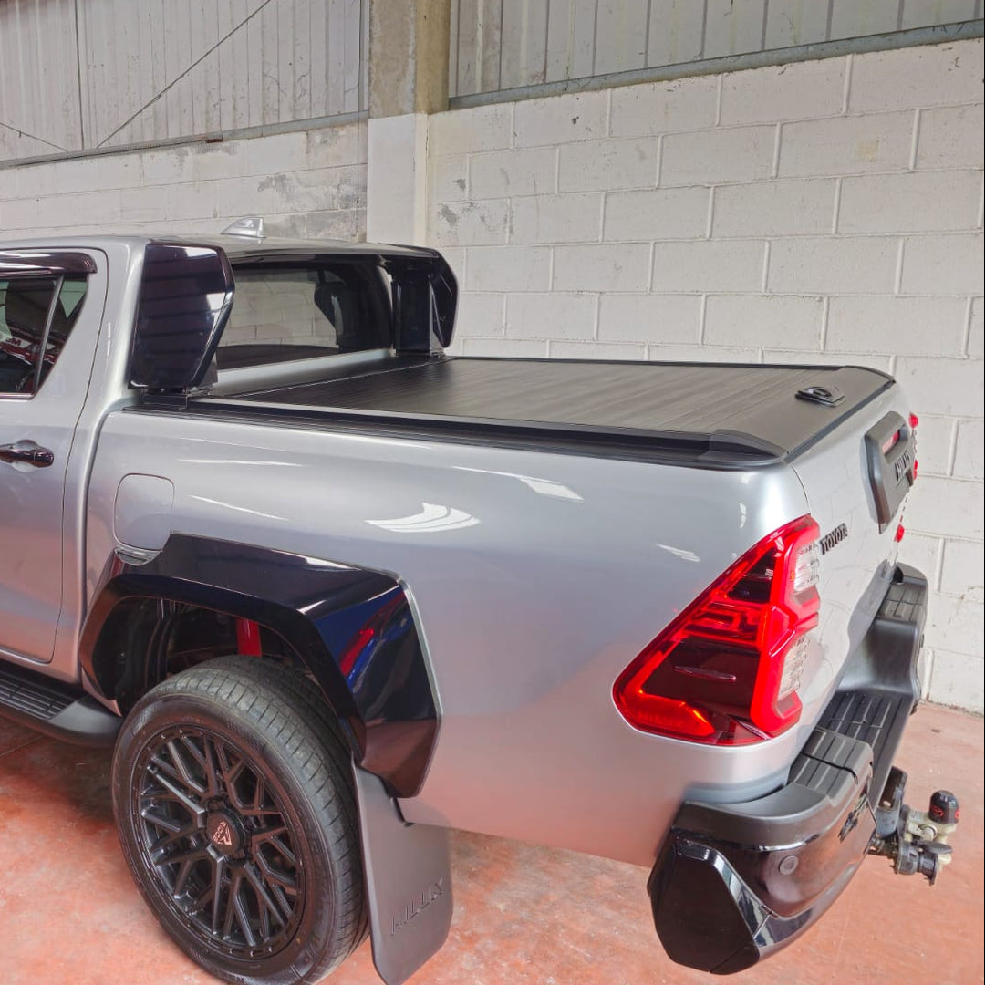 A silver Toyota Hilux pickup truck with black custom body kits, flared wheels, and a black tonneau cover, parked inside.
