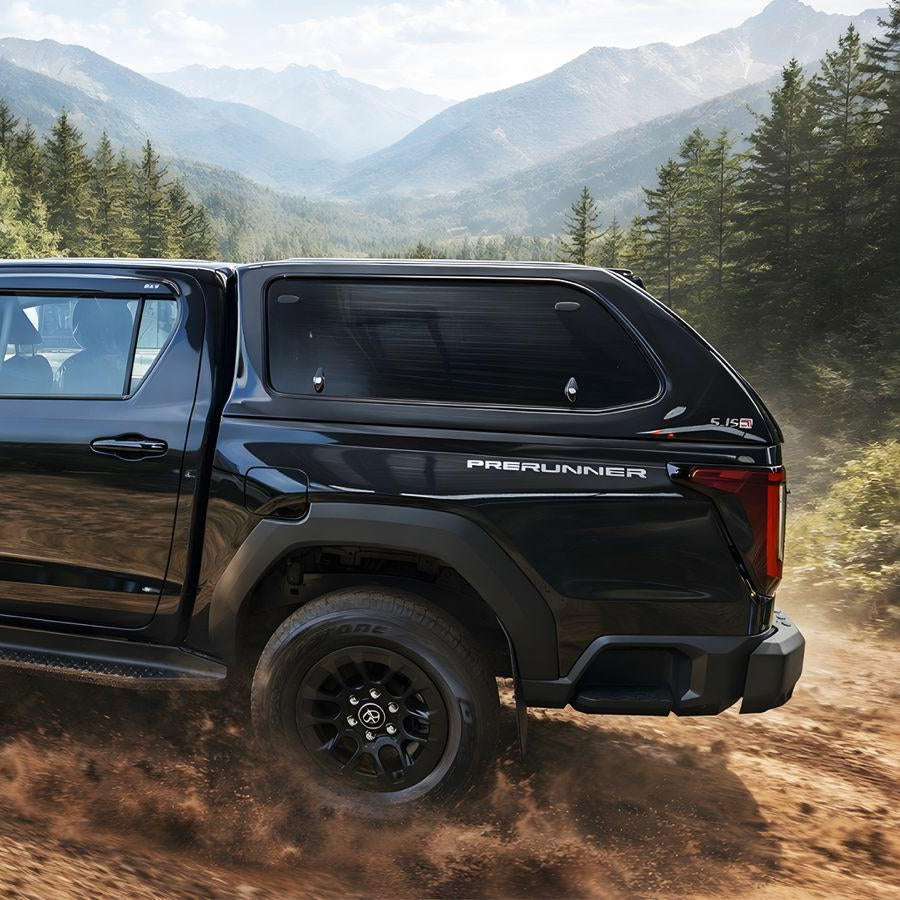 A black Ram 112 PreRunner pickup truck with a hard cap is shown driving on a dirt trail surrounded by a forest and mountains.