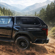 A black Ram 112 PreRunner pickup truck with a hard cap is shown driving on a dirt trail surrounded by a forest and mountains.